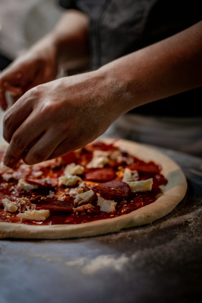Hands of a chef skillfully preparing a pepperoni pizza with fresh ingredients in a kitchen setting.